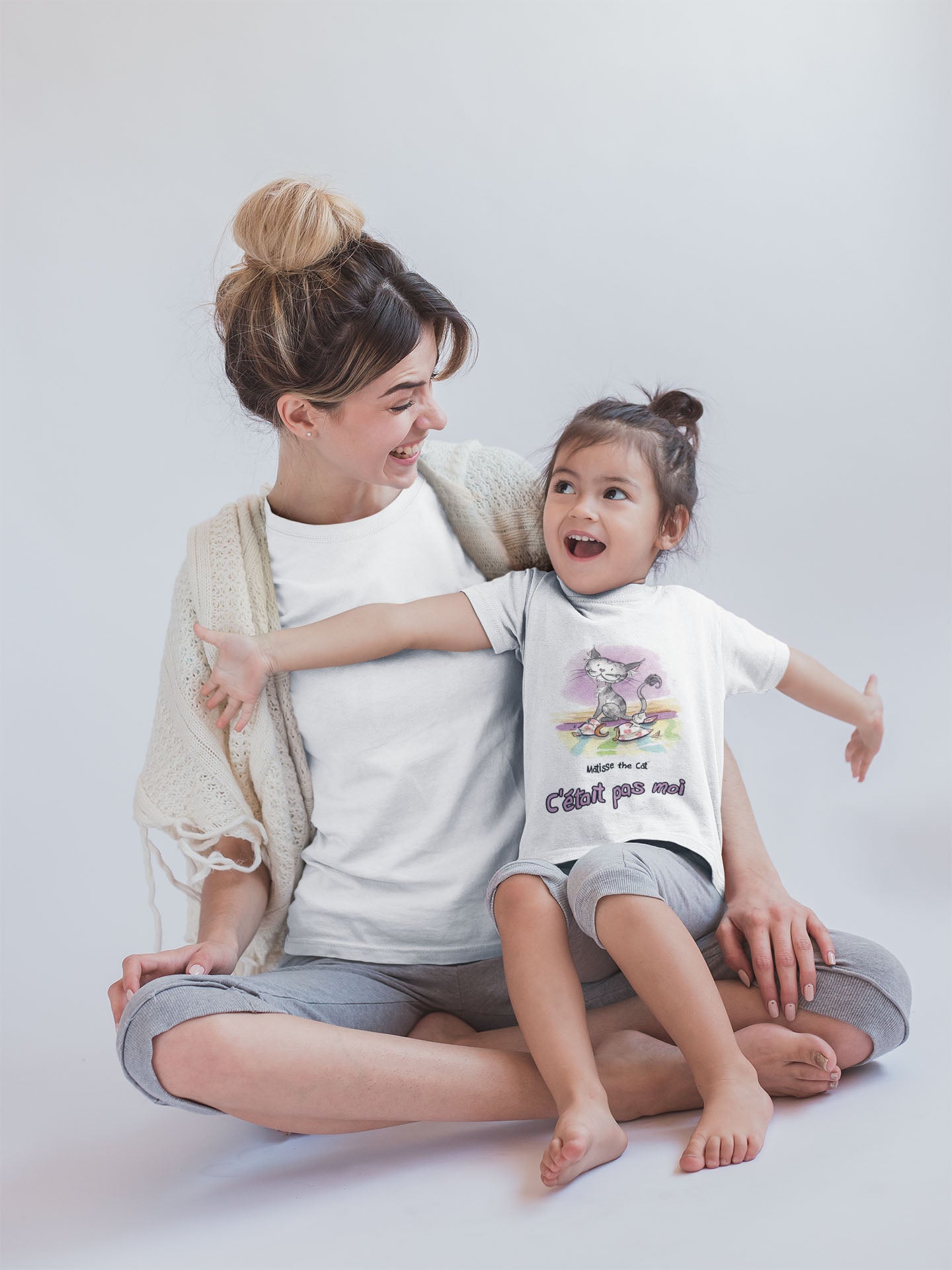 A white official, Matisse the Cat children’s t-shirt with the slogan “c'était pas moi” (It wasn’t me) featuring Matisse sitting on a vibrant carpet, his innocent gaze fixed upon a broken vase. Worn by a young girl sitting with her mum having fun.