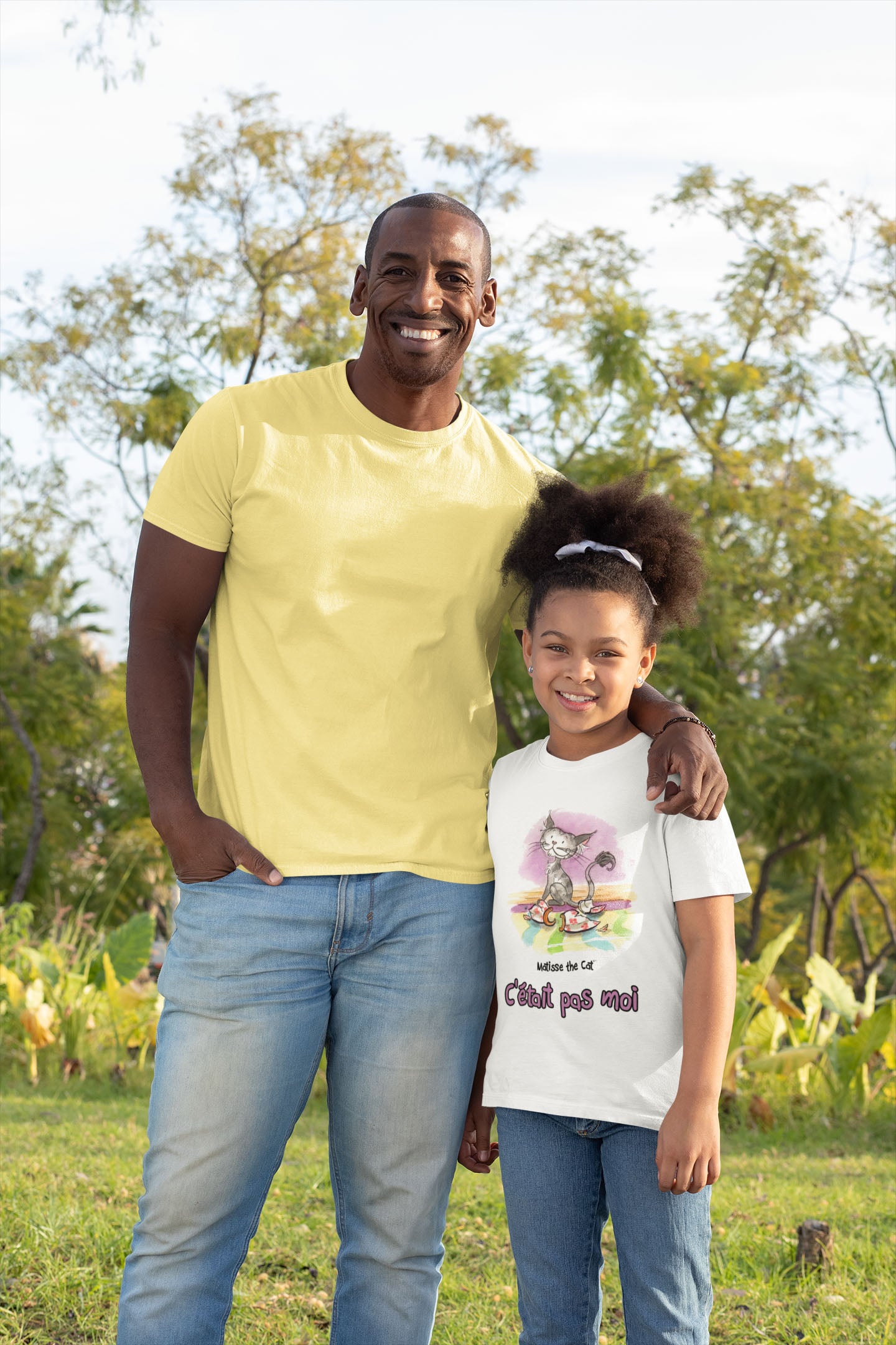 A white official, Matisse the Cat children’s t-shirt with the slogan “c'était pas moi” (It wasn’t me) featuring Matisse sitting on a vibrant carpet, his innocent gaze fixed upon a broken vase. Worn by a young girl standing beside her dad smiling.