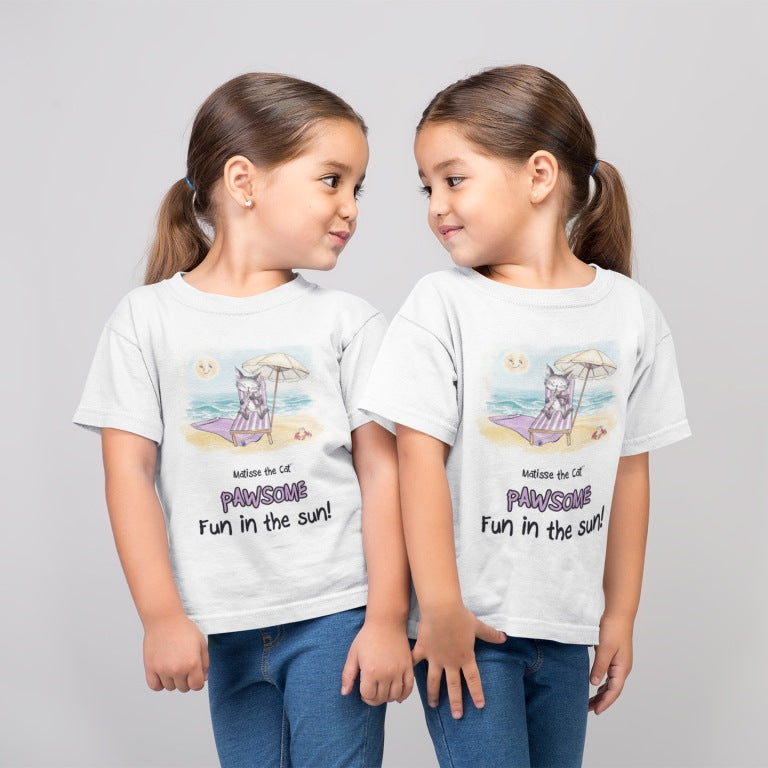 A white official, Matisse the Cat children’s t-shirt with the slogan ‘Pawsome Fun in the Sun ‘ showcases Matisse at the beach, relaxing on a sun lounger sipping a mile shake. Worn by twin girls, standing side by side looking at each other.