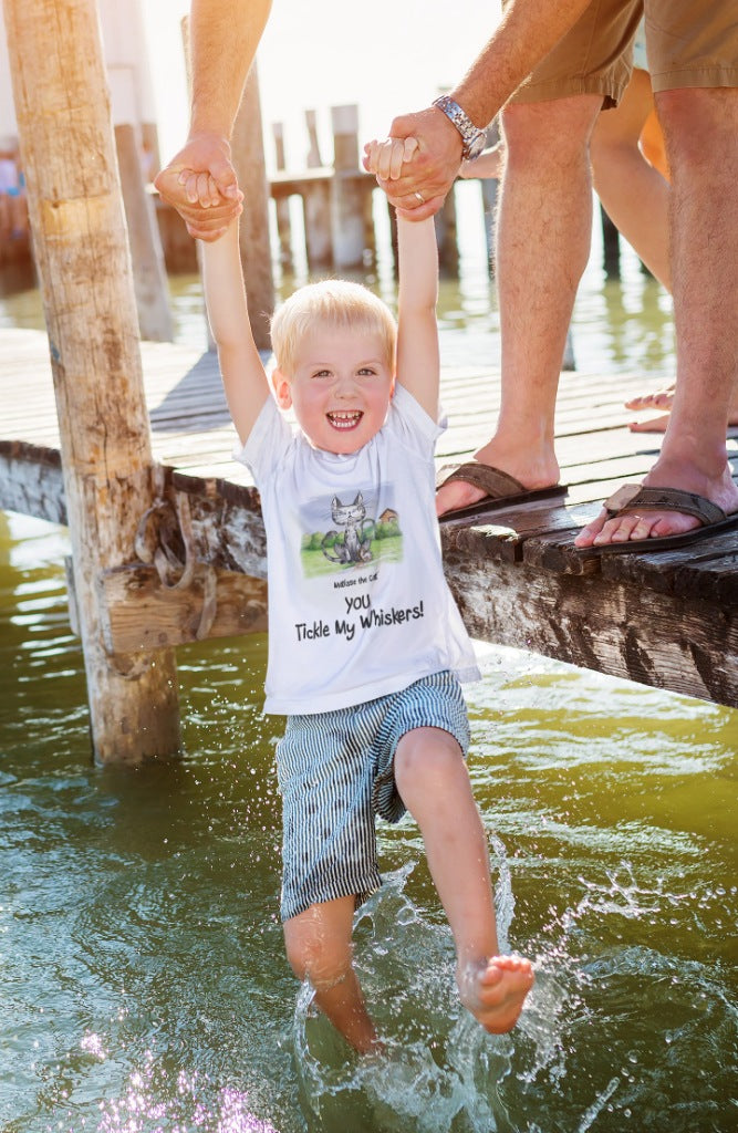 A white official, Matisse the Cat children’s t-shirt with the slogan ‘YOU Tickle My Whiskers ‘ showcases Matisse sitting in his garden smiling with a sleeping hedgehog curled up beside him resting on his leg. Worn by a young boy, dipping his feet in a lake being held by his father.