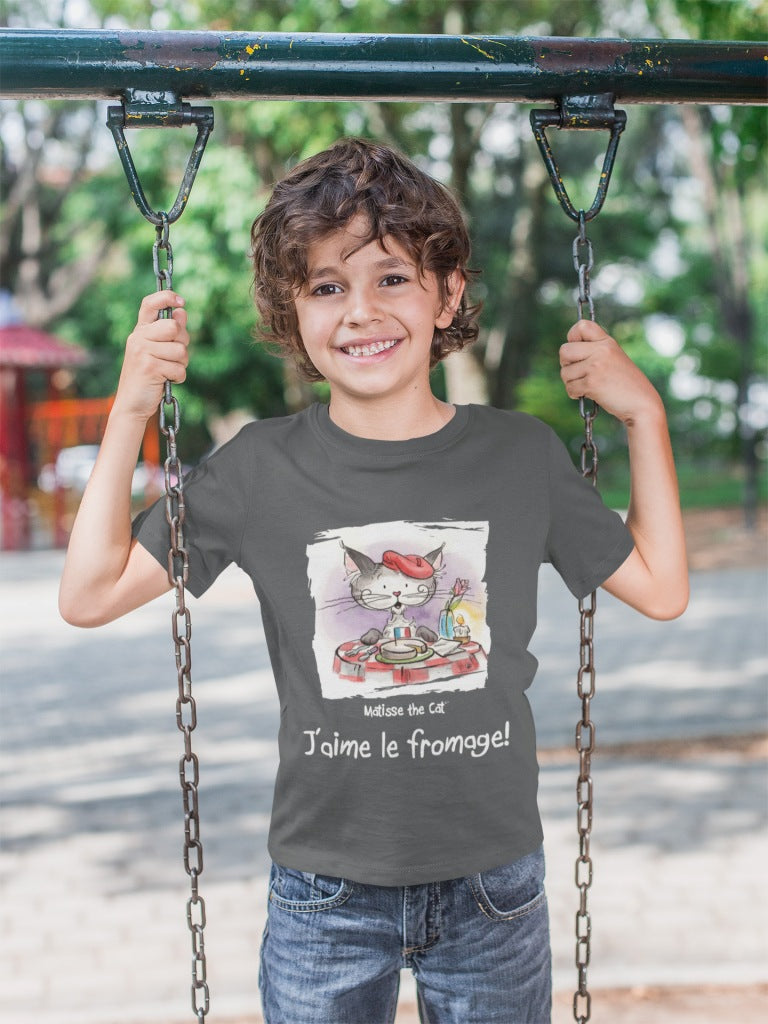 A asphalt grey official, Matisse the Cat children’s t-shirt with the slogan ‘Jaime le fromage ‘ showcases Matisse sitting at a French bistro table preparing to eat cheese. Worn by a young boy on a swing.