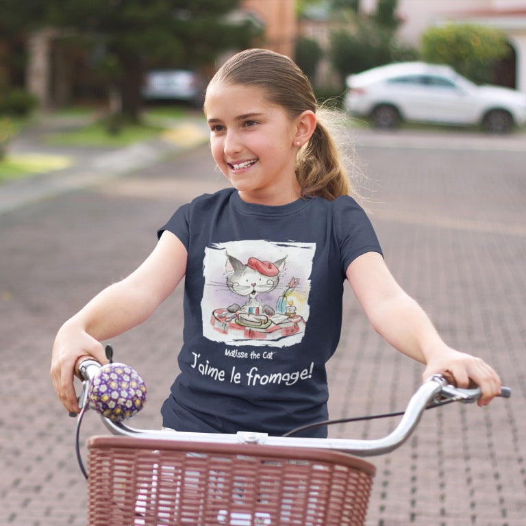 A navy blue official, Matisse the Cat children’s t-shirt with the slogan ‘Jaime le fromage ‘ showcases Matisse sitting at a French bistro table preparing to eat cheese. Worn by a young girl riding a bicycle.