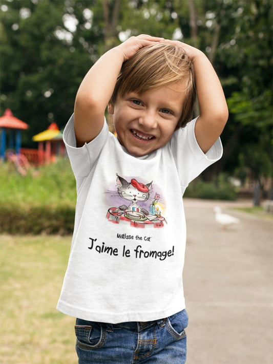 A white official, Matisse the Cat children’s t-shirt with the slogan ‘Jaime le fromage ‘ showcases Matisse sitting at a French bistro table preparing to eat cheese. Worn by a young boy in a park smiling and holding his hands on his head.