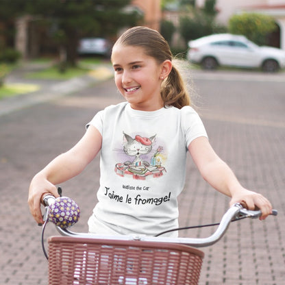 A white official, Matisse the Cat children’s t-shirt with the slogan ‘Jaime le fromage ‘ showcases Matisse sitting at a French bistro table preparing to eat cheese. Worn by a young girl riding a bicycle.