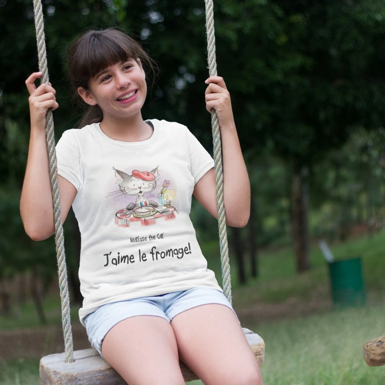 A white official, Matisse the Cat children’s t-shirt with the slogan ‘Jaime le fromage ‘ showcases Matisse sitting at a French bistro table preparing to eat cheese. Worn by a young girl sitting on a swing.