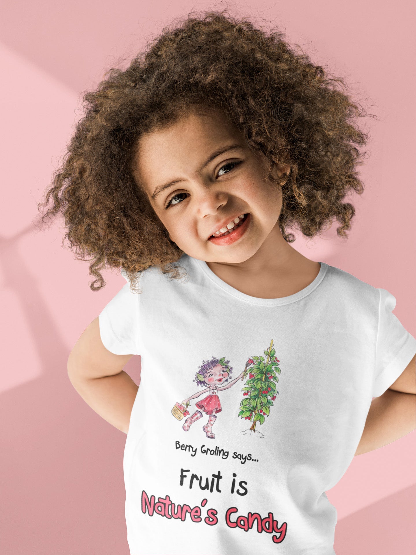 A white official Grolings kids' t-shirt, featuring the phrase 'Berry Groling says... Fruit is Nature’s Candy'. The t-shirt showcases Berry Groling, painting sweetness into raspberries on a bush. Berry Groling is holding a basket filled with fresh raspberries, capturing the abundance and delectable sweetness of nature's bounty. The scene encourages the wearer to appreciate and enjoy the natural delights provided by the Earth. Worn by a young girl smiling and standing in front of a pink background.