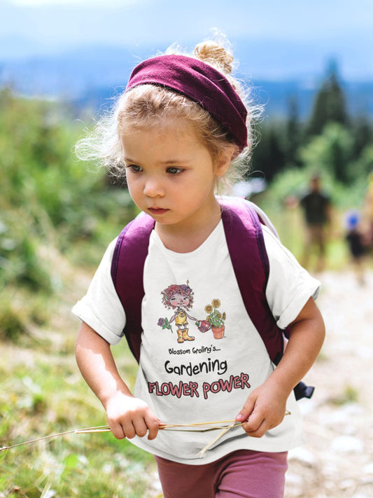A white official USA Grolings kids' t-shirt, featuring the phrase 'Blossom Groling... Gardening Flower Power’. The t-shirt showcases Blossom Groling, a character from ‘Matisse and the Topsy-Turvy Farm,’ watering a sunflower plant in a pot. Blossom is holding a beautiful flower in her other hand. The scene encourages the wearer to embrace a positive outlook and to share kindness and optimism, much like the blossoming flowers. Worn by a little girl on a hike through the countryside.