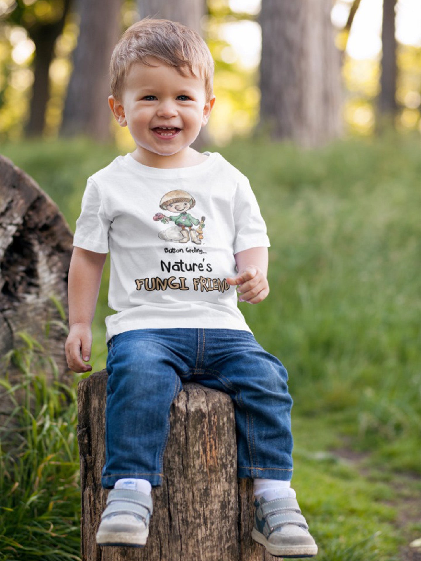 A white official Grolings kids' t-shirt, featuring the phrase 'Button Groling... Nature’s Fungi Friend.' The t-shirt showcases Button Groling, holding a little group of toadstools in one hand and a mushroom brush in the other. Beside Button Groling, there are two large puffball mushrooms, symbolising the importance of fungi in the forest ecosystem. Worn by a little boy sitting on a tree stump in a forest.