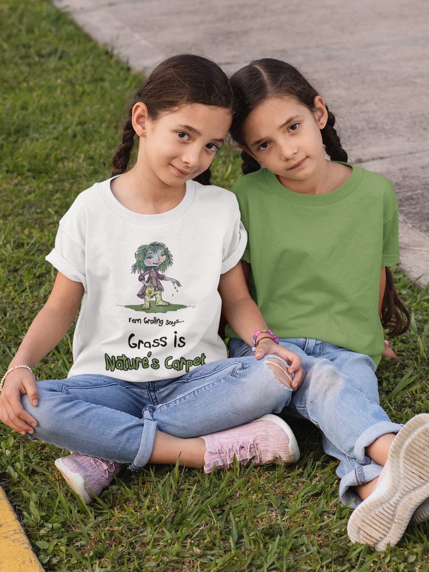 A white official Grolings kids' t-shirt, featuring the phrase 'Fern Groling says... Grass is Nature’s Carpet.' The t-shirt showcases Fern Groling, standing on green ground and planting grass seeds. The scene emphasises the importance of grass as a natural carpet, symbolising the lushness and vitality it brings to the environment. Worn by a twin girl sitting on a grass path with her sister.