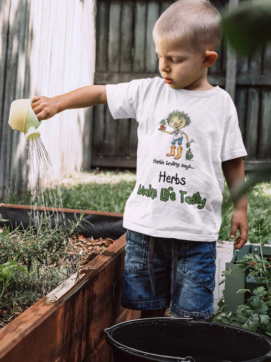 A white official Grolings kids' t-shirt, featuring the phrase 'Herbie Groling says... Herbs Make Life Tasty. The t-shirt showcases Herbie Groling, holding a basil plant in a pot. Beside Herbie, a thriving rosemary bush adds to the scene. The imagery highlights the delightful flavours and aromatic qualities that herbs bring to our lives, encouraging an appreciation for the culinary joys and benefits of herbs. Worn by a young boy watering a rosemary bush in a greenhouse.