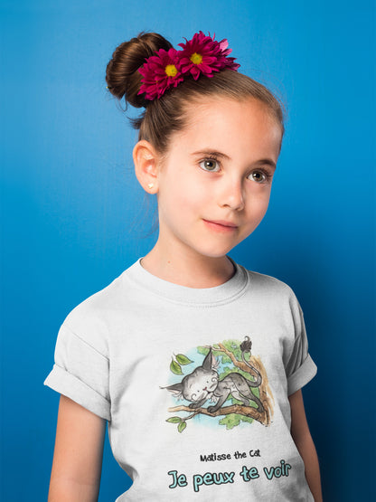 A white official, Matisse the Cat children’s t-shirt with the slogan ‘Je peux te voir’ showcases Matisse sitting in his garden smiling with a sleeping hedgehog curled up beside him resting on his leg. Worn by a girl posing in front of a blue background with a flower in her hair.