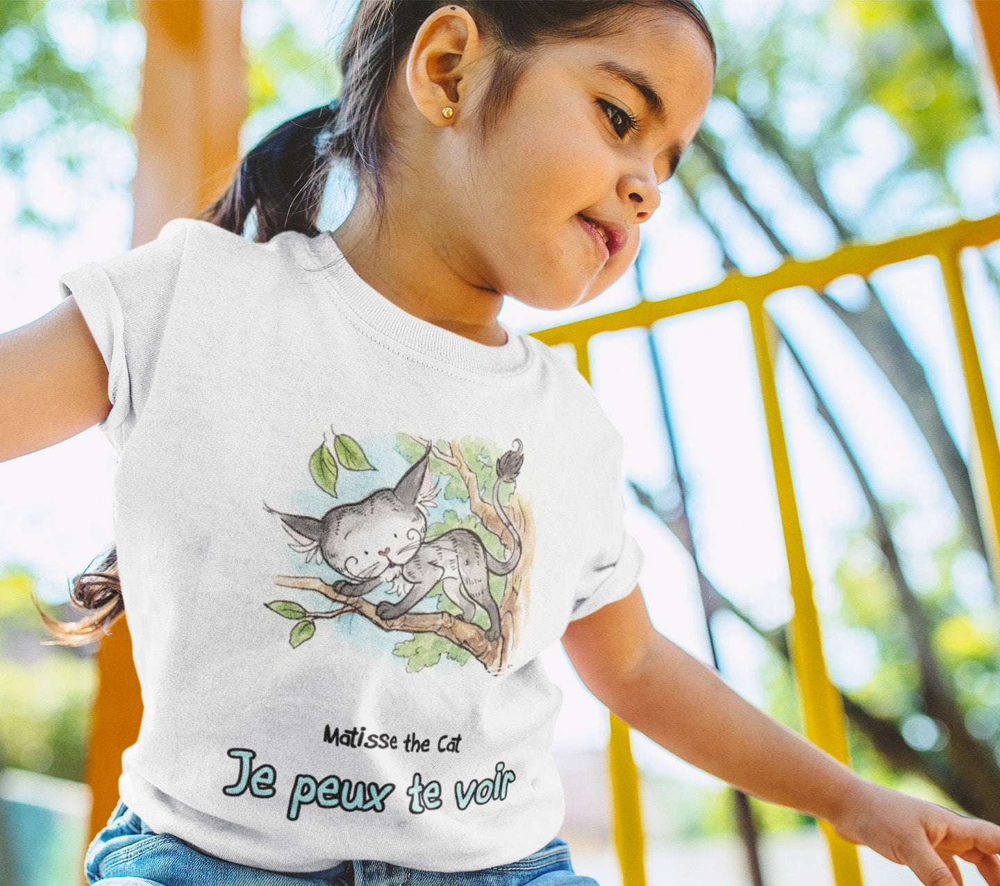 A white official, Matisse the Cat children’s t-shirt with the slogan ‘Je peux te voir’ showcases Matisse sitting in his garden smiling with a sleeping hedgehog curled up beside him resting on his leg. Worn by a little girl playing on a climbing frame.
