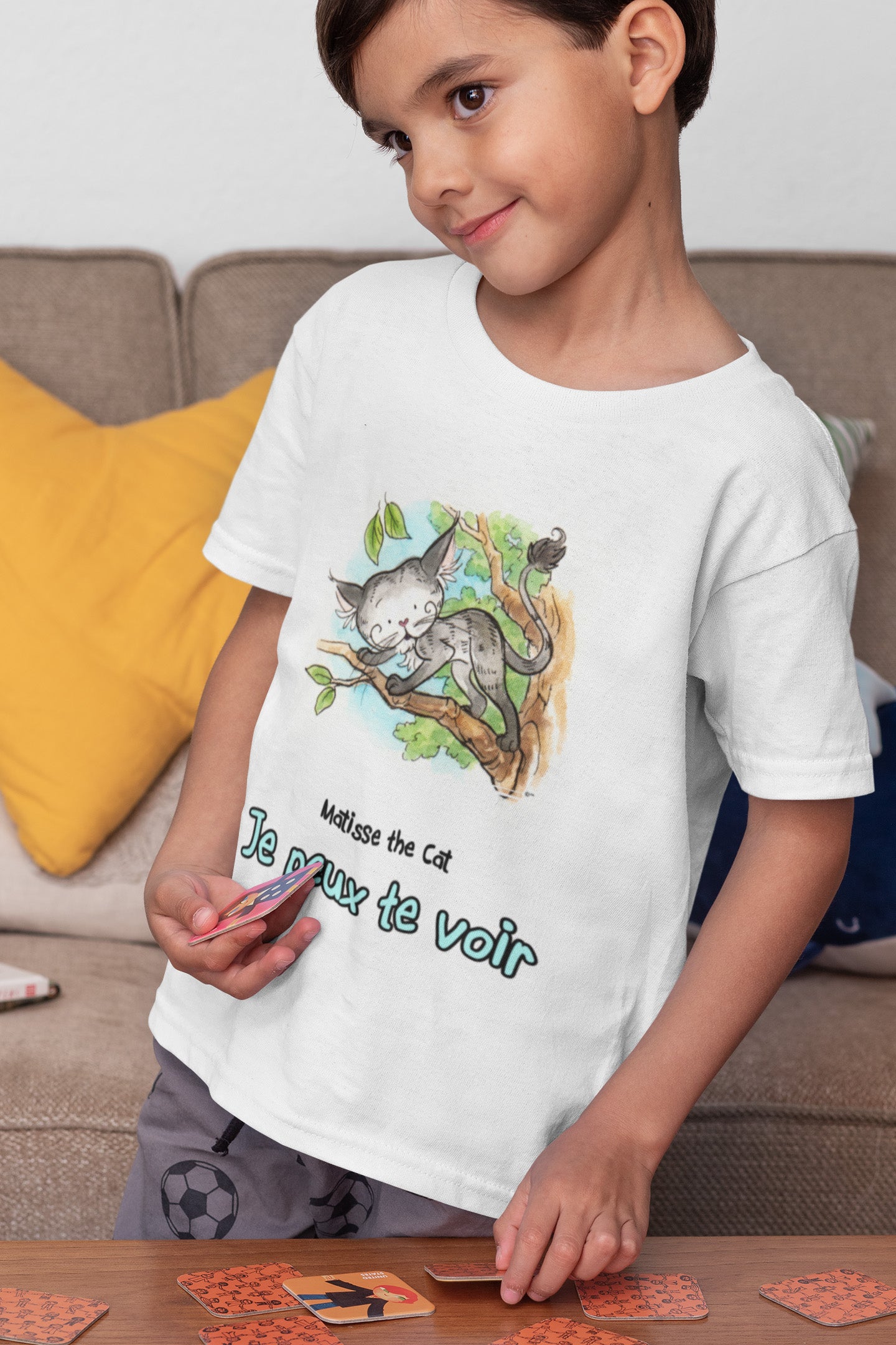 A white official, Matisse the Cat children’s t-shirt with the slogan ‘Je peux te voir’ showcases Matisse sitting in his garden smiling with a sleeping hedgehog curled up beside him resting on his leg. Worn by a young boy playing a card game in his living room.