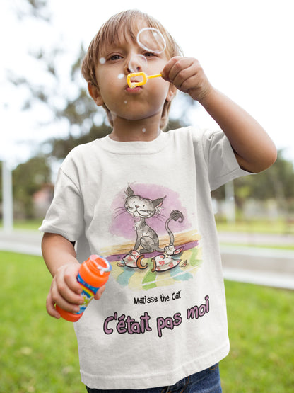 A white official, Matisse the Cat children’s t-shirt with the slogan “c'était pas moi” (It wasn’t me) featuring Matisse sitting on a vibrant carpet, his innocent gaze fixed upon a broken vase. Worn by a young boy making bubbles.