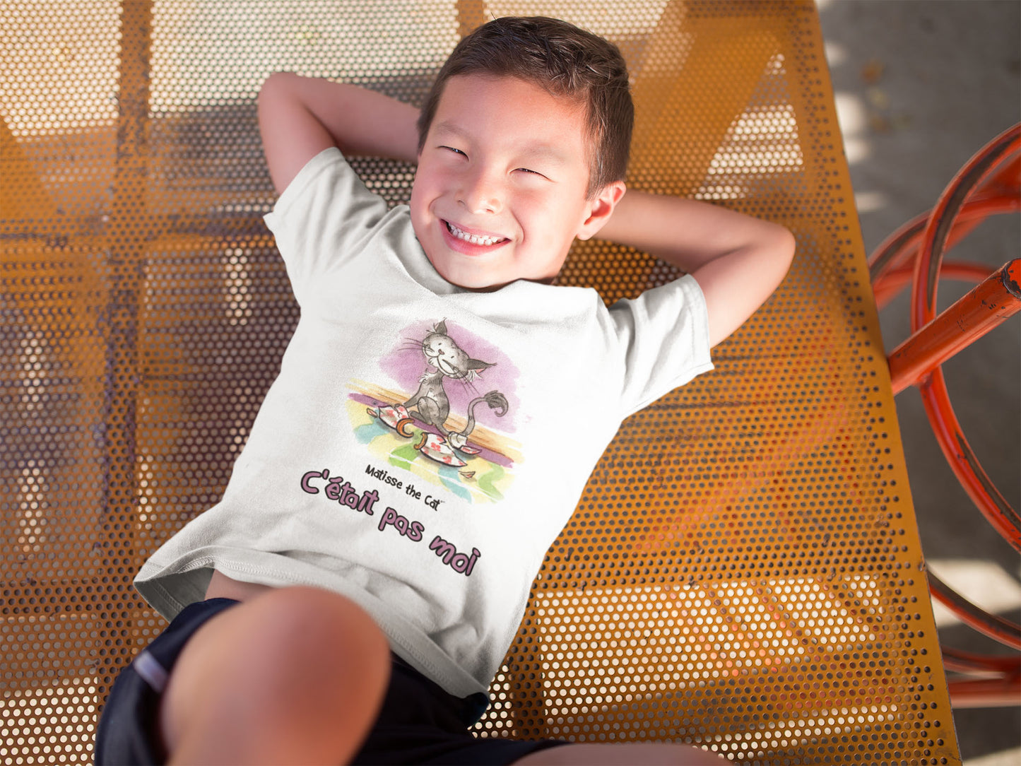 A white official, Matisse the Cat children’s t-shirt with the slogan “c'était pas moi” (It wasn’t me) featuring Matisse sitting on a vibrant carpet, his innocent gaze fixed upon a broken vase. Worn by a happy young boy in a playground.