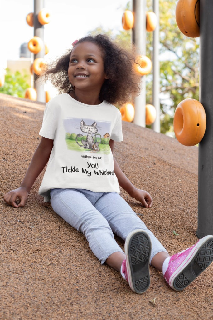 A white official, Matisse the Cat children’s t-shirt with the slogan ‘YOU Tickle My Whiskers ‘ showcases Matisse sitting in his garden smiling with a sleeping hedgehog curled up beside him resting on his leg. Worn by a young girl sitting on a hill in a play park.