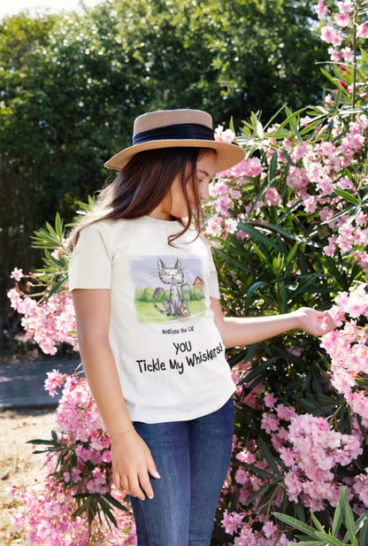 A white official, Matisse the Cat children’s t-shirt with the slogan ‘YOU Tickle My Whiskers ‘ showcases Matisse sitting in his garden smiling with a sleeping hedgehog curled up beside him resting on his leg. Worn by a young girl wearing a hat gently touching flower petals on a bush.