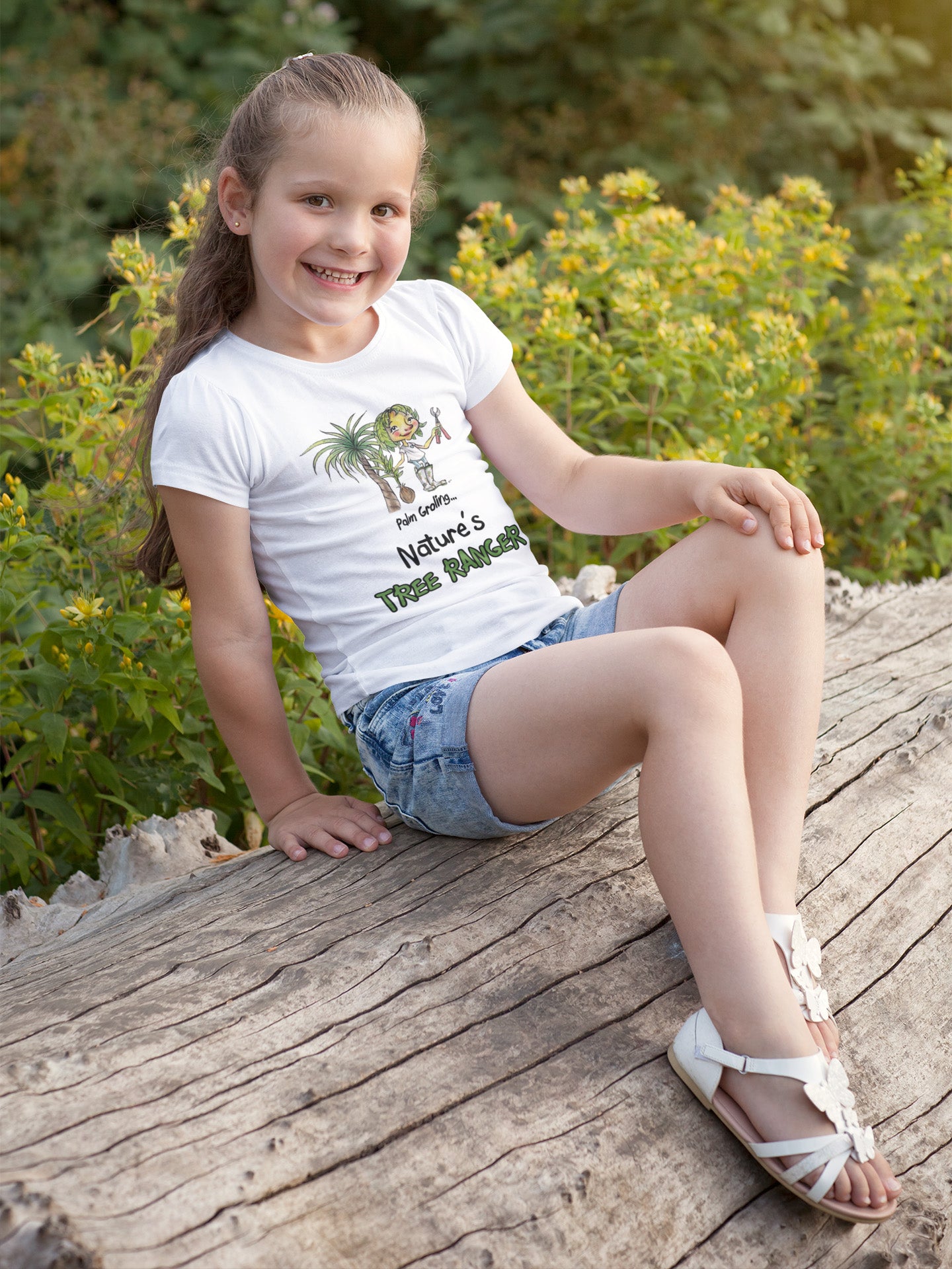 A white official USA Grolings kids' t-shirt, featuring the phrase 'Palm Groling... Nature’s Tree Ranger.' The t-shirt showcases Palm Groling, holding a young tree in one hand and pruning shears in the other. In the background, a majestic palm tree stands tall. The scene emphasises the importance and grandeur of trees as essential components of the Earth's ecosystem, promoting a sense of awe and reverence for these green giants. Worn by a young girl sitting on a tree trunk.